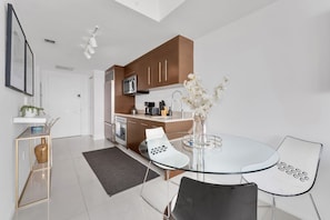 A modern kitchen with brown cabinets, a white countertop, and a dining area with a glass table and chairs.