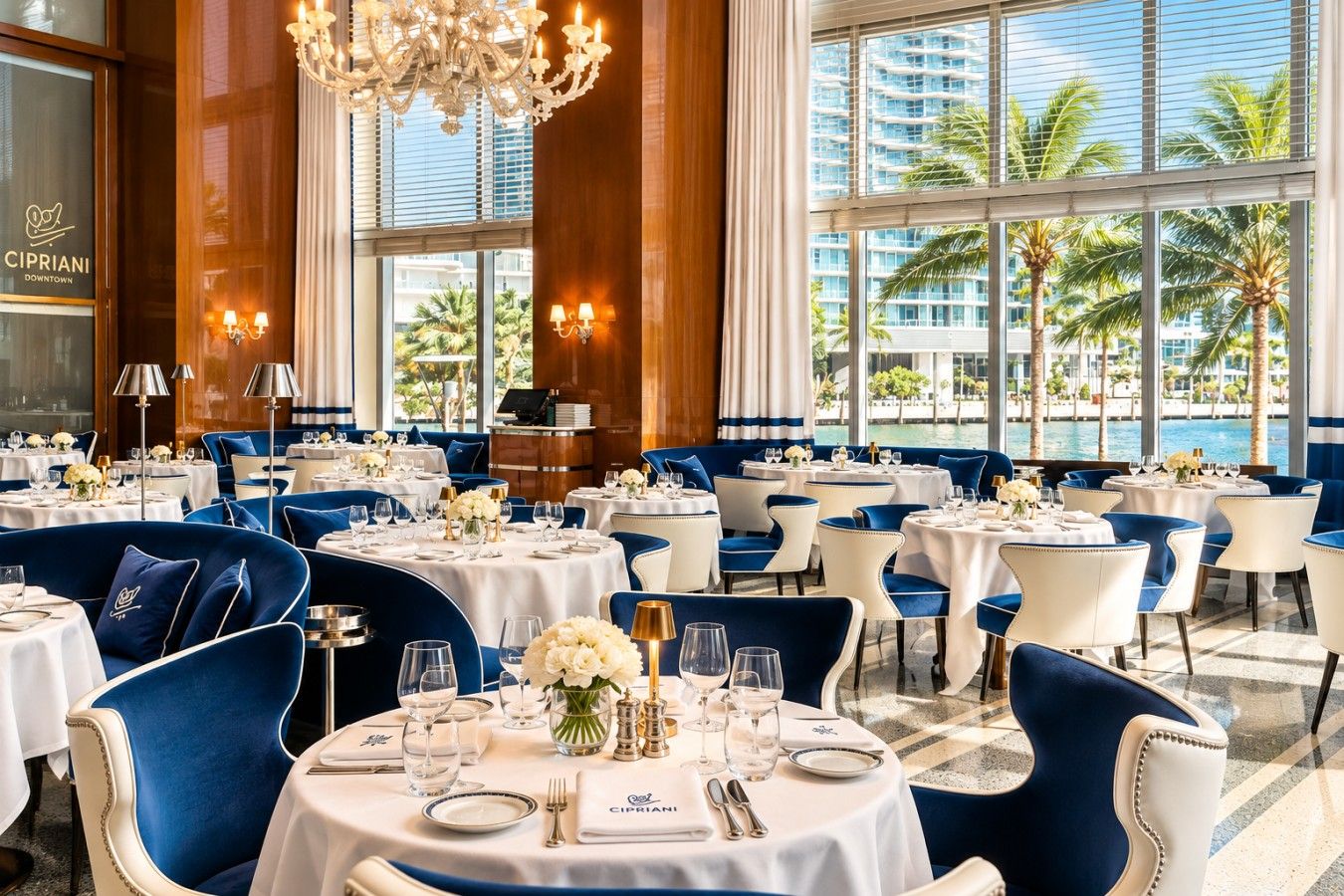 A restaurant with round tables, blue upholstered chairs, and a view of palm trees and buildings through large windows.