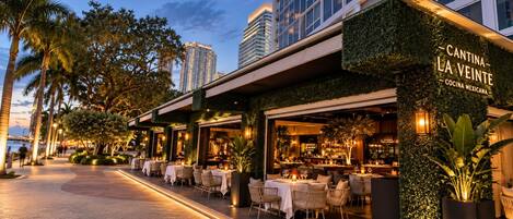 Un área de comedor al aire libre con mesas y sillas, un edificio con pared verde y un letrero que dice 'CANTINA LA VEINTE COCINA MEXICANA'.