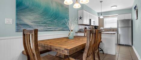 A dining area with a wooden table and chairs, a large wave painting on the wall, and a kitchen in the background.