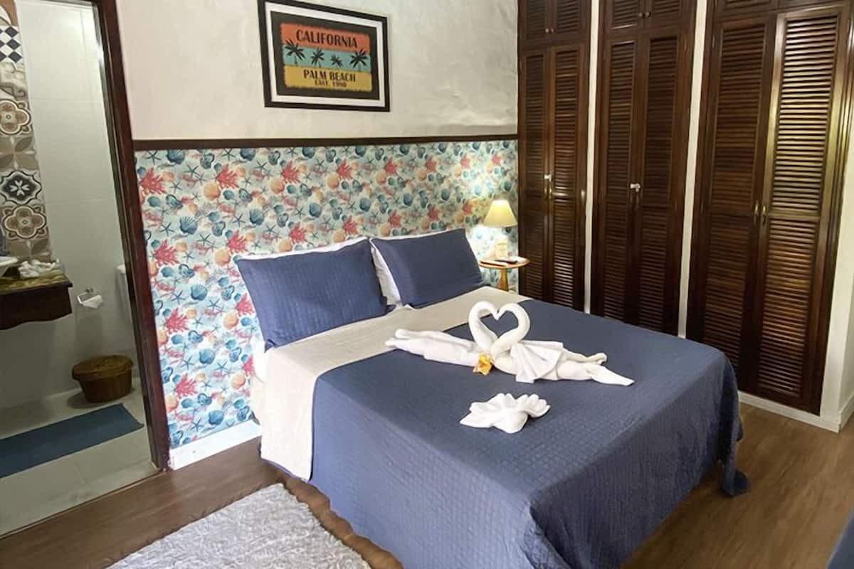 Un lit bien fait, avec une couvre-lit bleue et des oreillers, une serviette pliée en forme de cœur, un cadre décoratif accroché au mur et une armoire en bois.