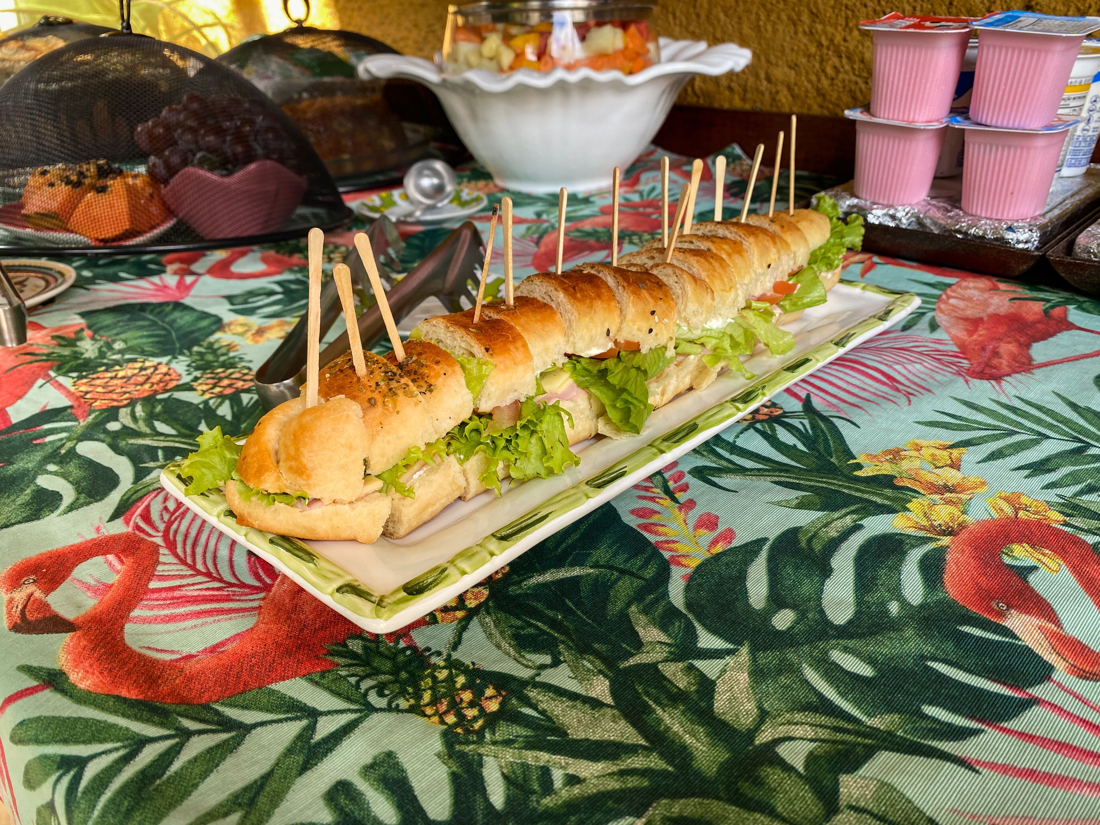 Een lang belegd broodje met satéprikkers op een bord, staand op een tafel met een bloemrijk tafelkleed.