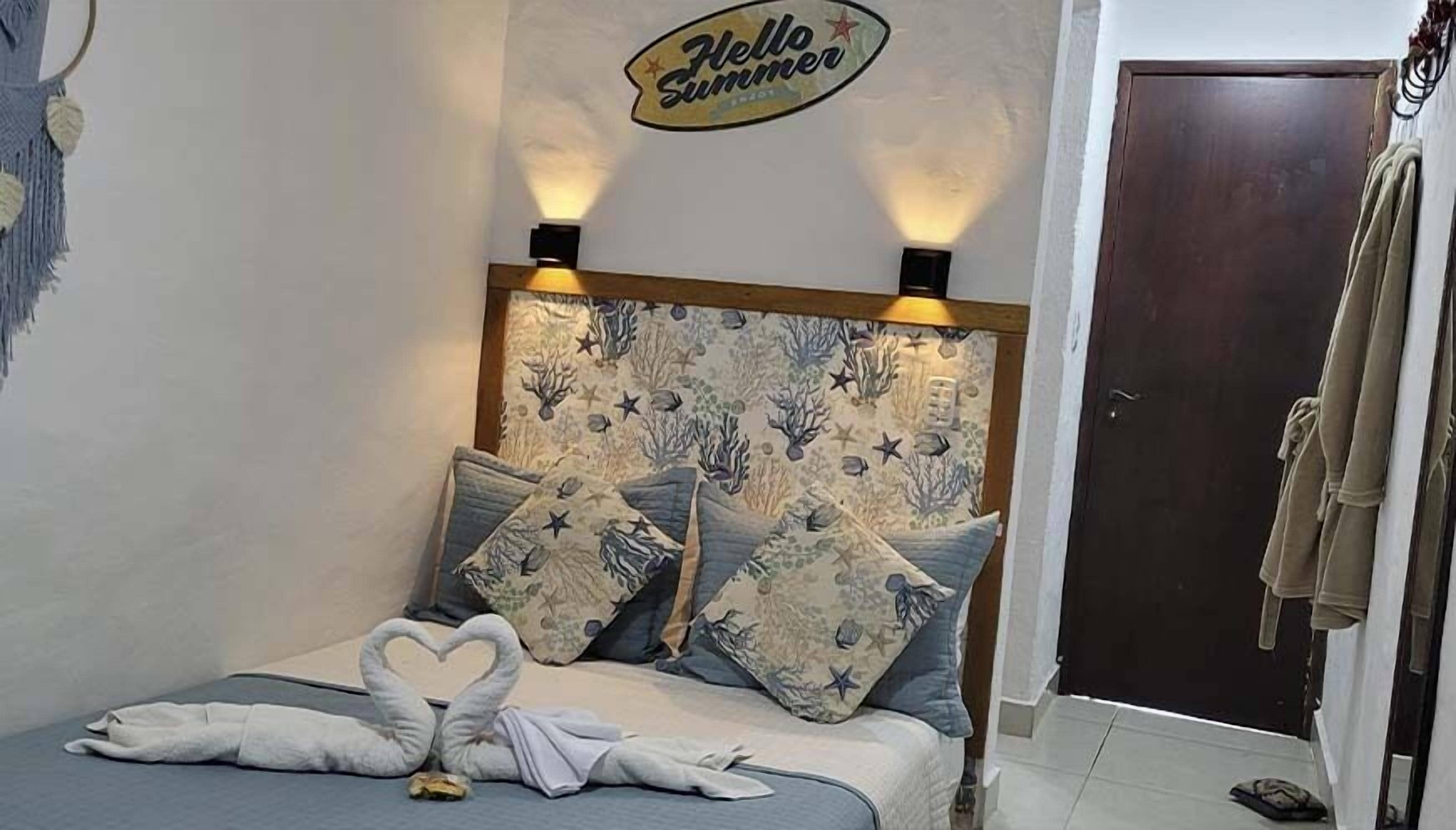 A neatly made bed with heart-shaped towels, a wall with a 'Hello Summer' sign, and a door to the right.