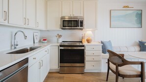 Une cuisine moderne avec des meubles blancs, des appareils en acier inoxydable, un coin repas avec une table blanche et des chaises en bois, et un tableau accroché au mur.