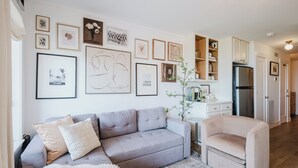 Un salon avec un canapé gris, un fauteuil beige et une table basse. On y trouve des tableaux encadrés au mur, un meuble blanc et un réfrigérateur.