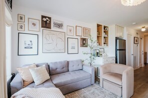 A living room with a grey sofa, a beige armchair, and a coffee table. There are framed pictures on the wall, a white cabinet, and a refrigerator.
