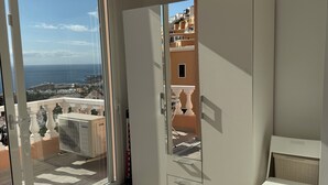 Een balkon met uitzicht op zee en gebouwen, een schuifdeur, een witte garderobe en een kleine witte tafel.