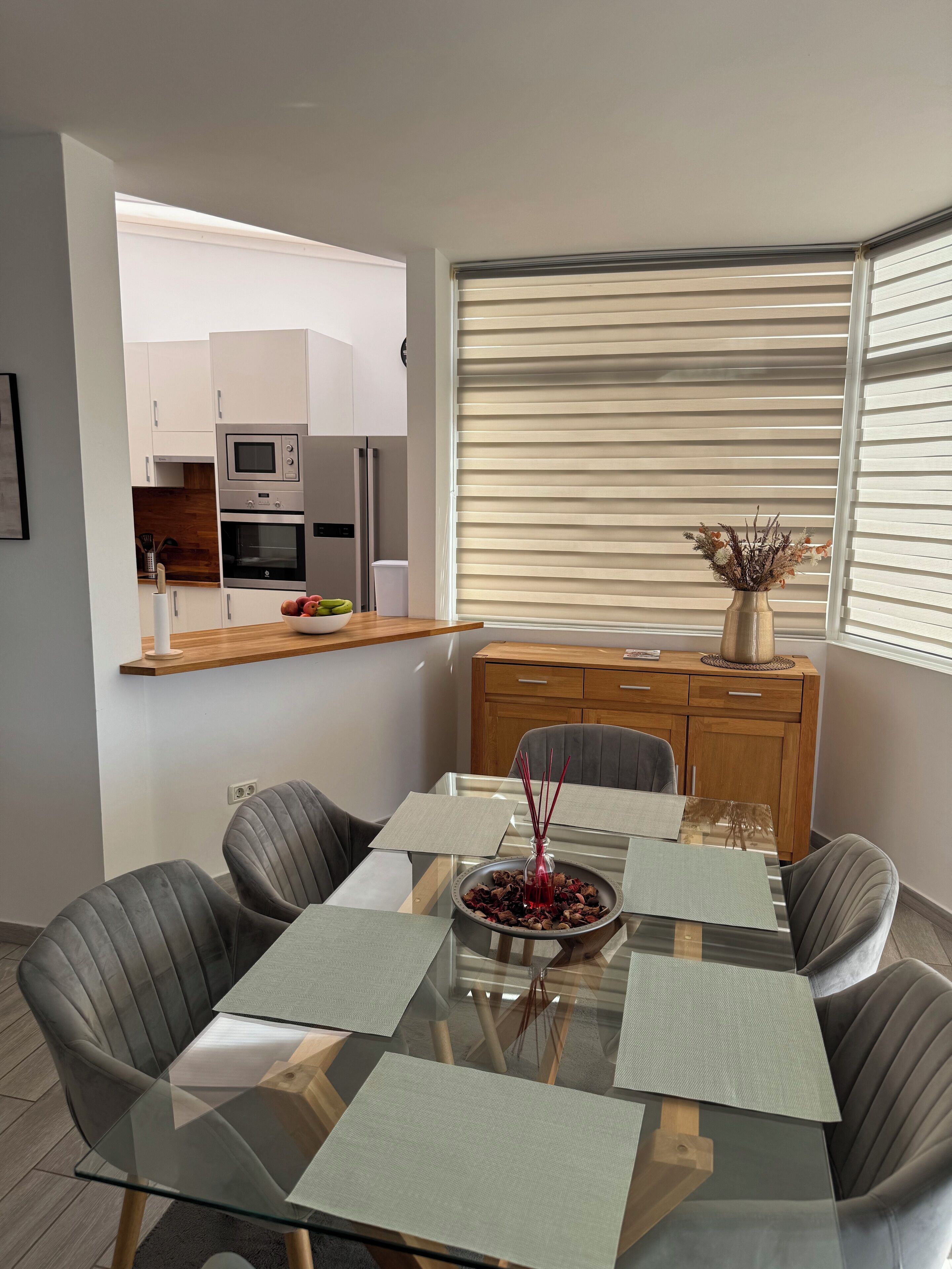 A modern dining area with a glass table, grey upholstered chairs, and a wooden cabinet.