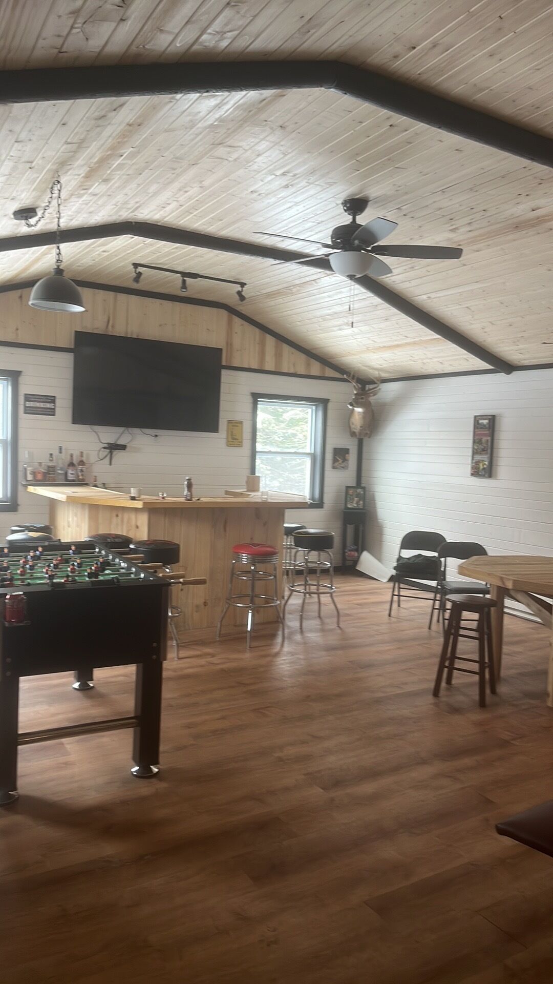 A spacious room with a wooden ceiling, a ceiling fan, a foosball table, bar stools, and a bar area.