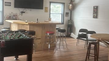 A spacious room with a wooden ceiling, a ceiling fan, a foosball table, bar stools, and a bar area.