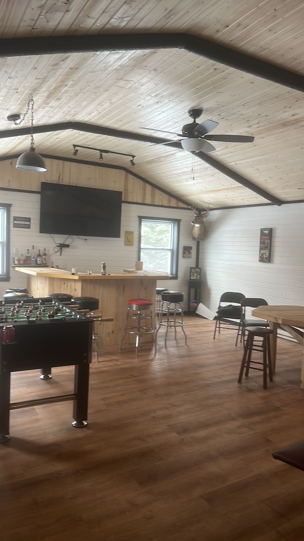 A spacious room with a wooden ceiling, a ceiling fan, a foosball table, bar stools, and a bar area.