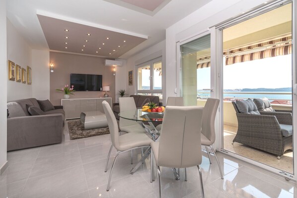 A modern living room with a glass dining table, grey sofas, and a balcony with a view of the sea.