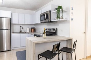 A modern kitchen with white cabinets, stainless steel appliances, a wooden island with two black chairs, and a wine bottle. - Beach Apartment in Río Grande (Villa Quenepa) (Rio Grande)
