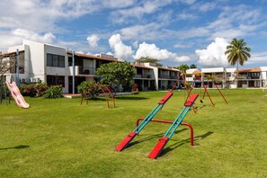 A playground with swings and a slide, surrounded by buildings with balconies and a clear sky.