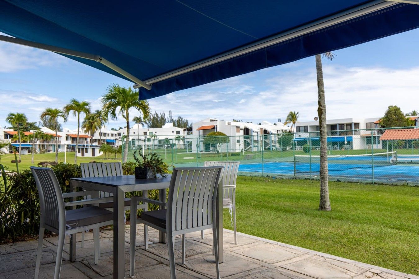 Un patio exterior con una mesa y sillas, un toldo azul, y una vista de una piscina y edificios al fondo.