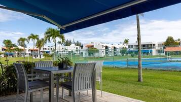 Un patio exterior con una mesa y sillas, un toldo azul, y una vista de una piscina y edificios al fondo.