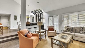Un salon moderne comprenant un canapé, un fauteuil, une table basse et un coin cuisine visible à travers une cloison en verre.