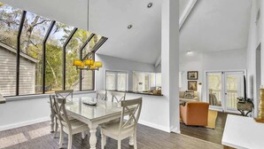 Un vaste espace à manger avec une table en verre et des chaises blanches, de grandes fenêtres équipées de stores et une vue sur l’extérieur.
