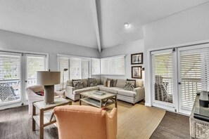 A living room with a sofa, coffee table, and armchair, large windows with blinds, and a door with a view. - Dream Getaway w/ wraparound deck on Hilton Head! (Hilton Head Island)
