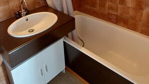 A bathroom with a white bathtub, a sink with a dark countertop, and brown tiled walls. - Bright 14m2 studio with unobstructed view (Saint-Michel-de-Chaillol)