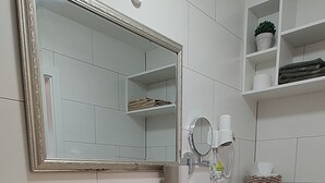 Ein modernes Badezimmer mit einem weißen Waschbecken, Spiegel und Toilette. Auf dem Boden liegt ein grünes Handtuch und neben der Toilette steht ein kleiner Mülleimer.