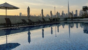 A pool with lounge chairs and umbrellas, a city skyline in the background, and a clear sky. - Luxury 1BDwith Stunning Burj Khalifa View- Self Check-in (Dubai)