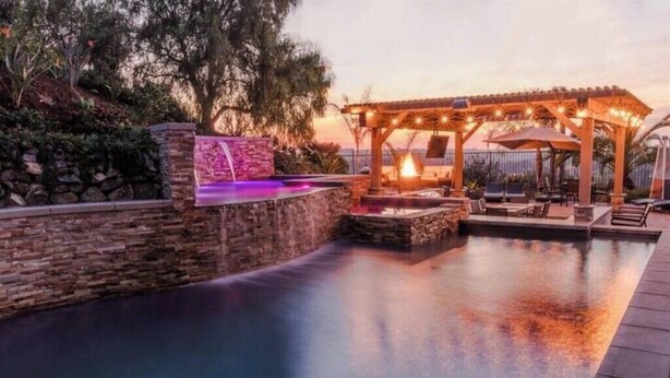 Un área de piscina con un muro de piedra, una pérgola con luces colgantes y sillones de descanso.