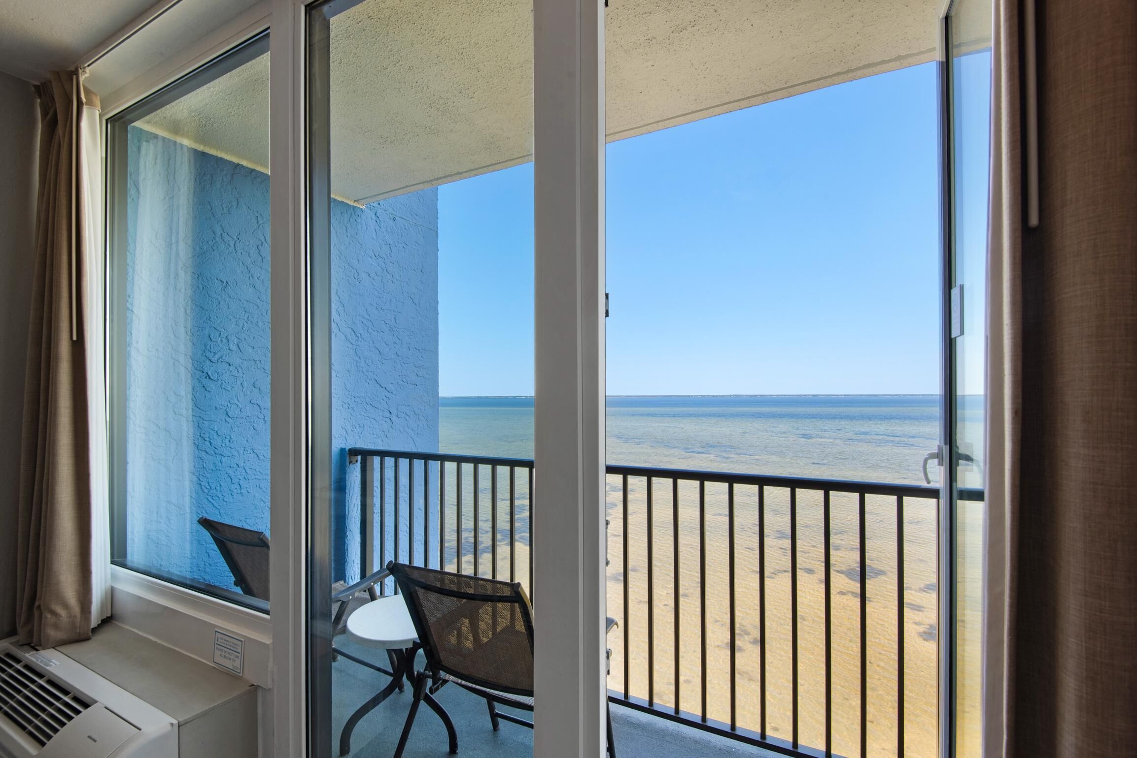 A balcony with a view of the beach, a chair, and a table.