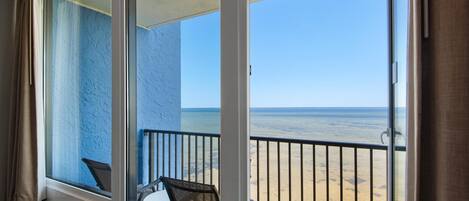 A balcony with a view of the beach, a chair, and a table.
