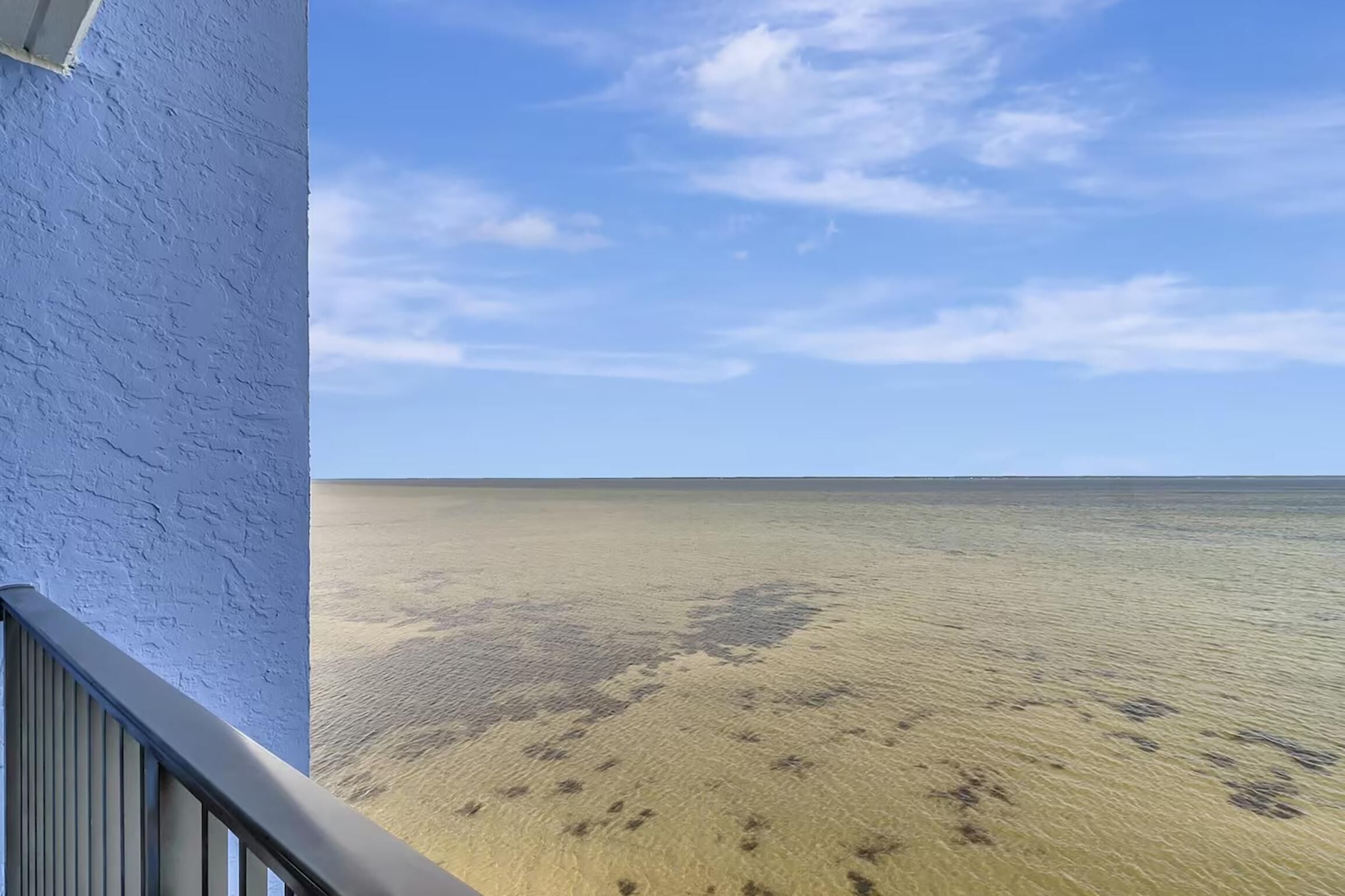Ein Balkon mit freiem Blick auf einen Sandstrand und das Meer unter blauem Himmel.
