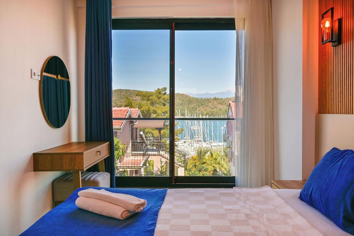 A hotel room with a bed, a desk, a mirror, and a view of a marina and hills.