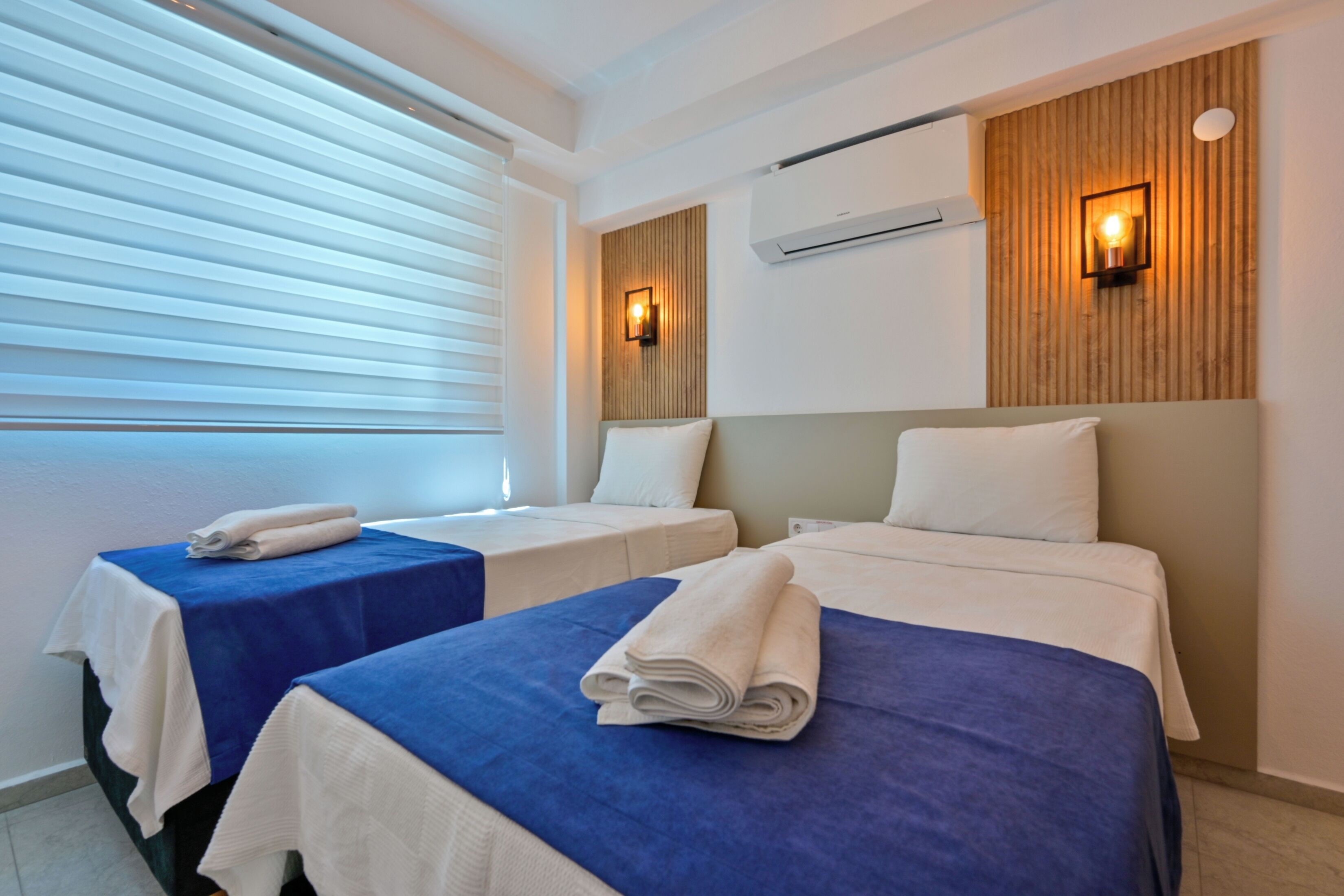 A hotel room with two beds, each with a blue and white bedspread, and a wooden panel wall with a wall-mounted air conditioner and two wall sconces.