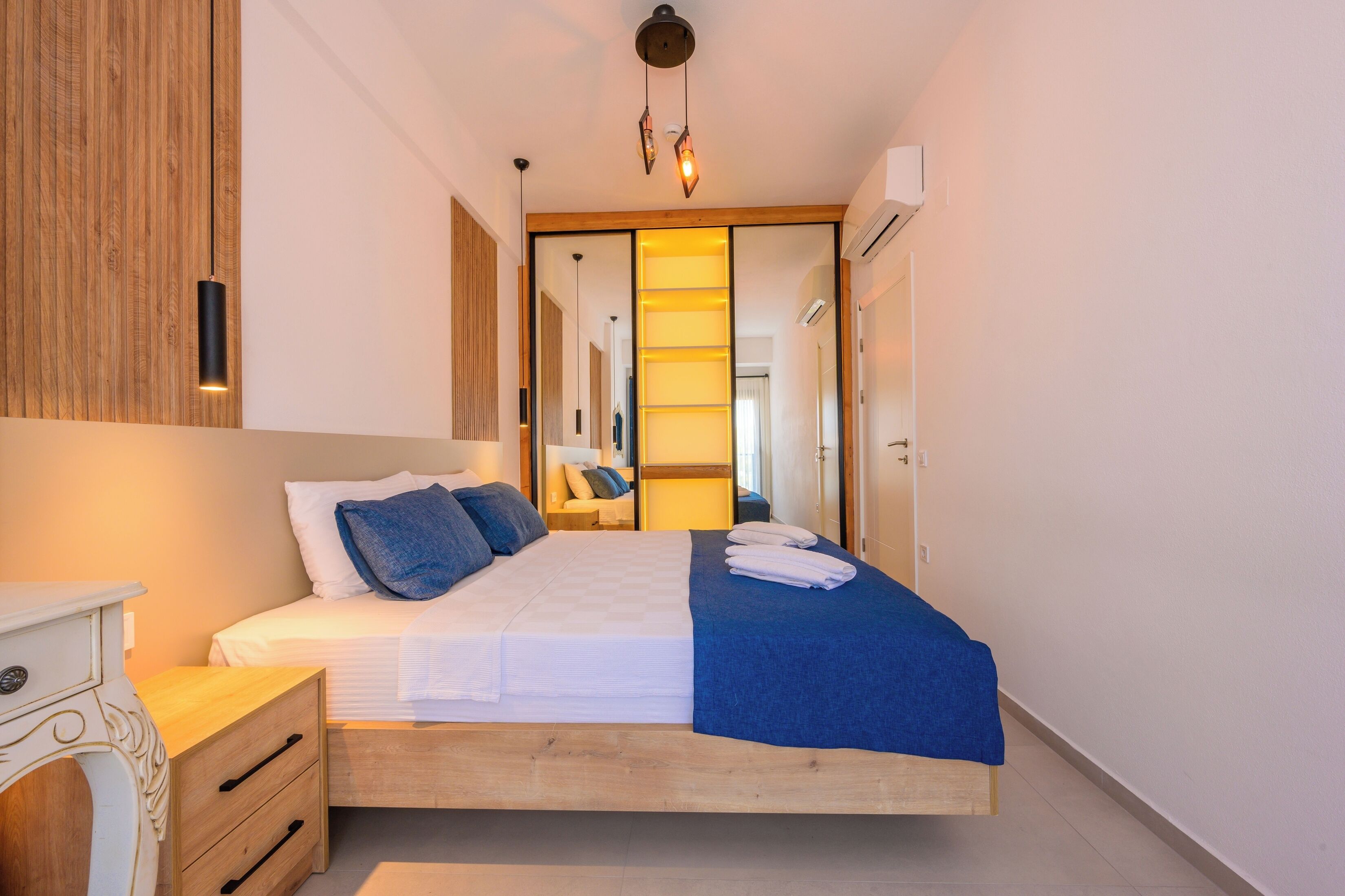 Ein modernes Schlafzimmer mit einem Holz-Kopfteil, einem weißen Bett mit blauer Bettwäsche, einem Nachttisch mit Schubladen und einer Hängeleuchte.