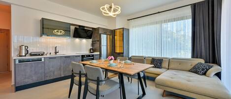 A modern living room with a sofa, dining table, and chairs. There is a kitchen area with a TV and a large window with curtains.