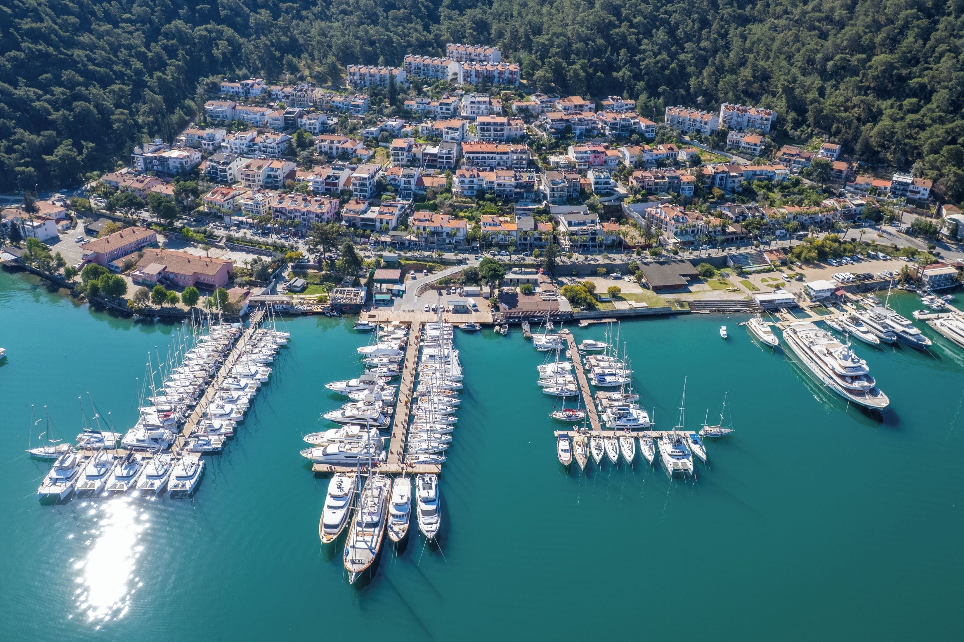Une vue aérienne d’une ville côtière, avec un port de plaisance rempli de bateaux, entourée de bâtiments résidentiels et d’une forêt dense.