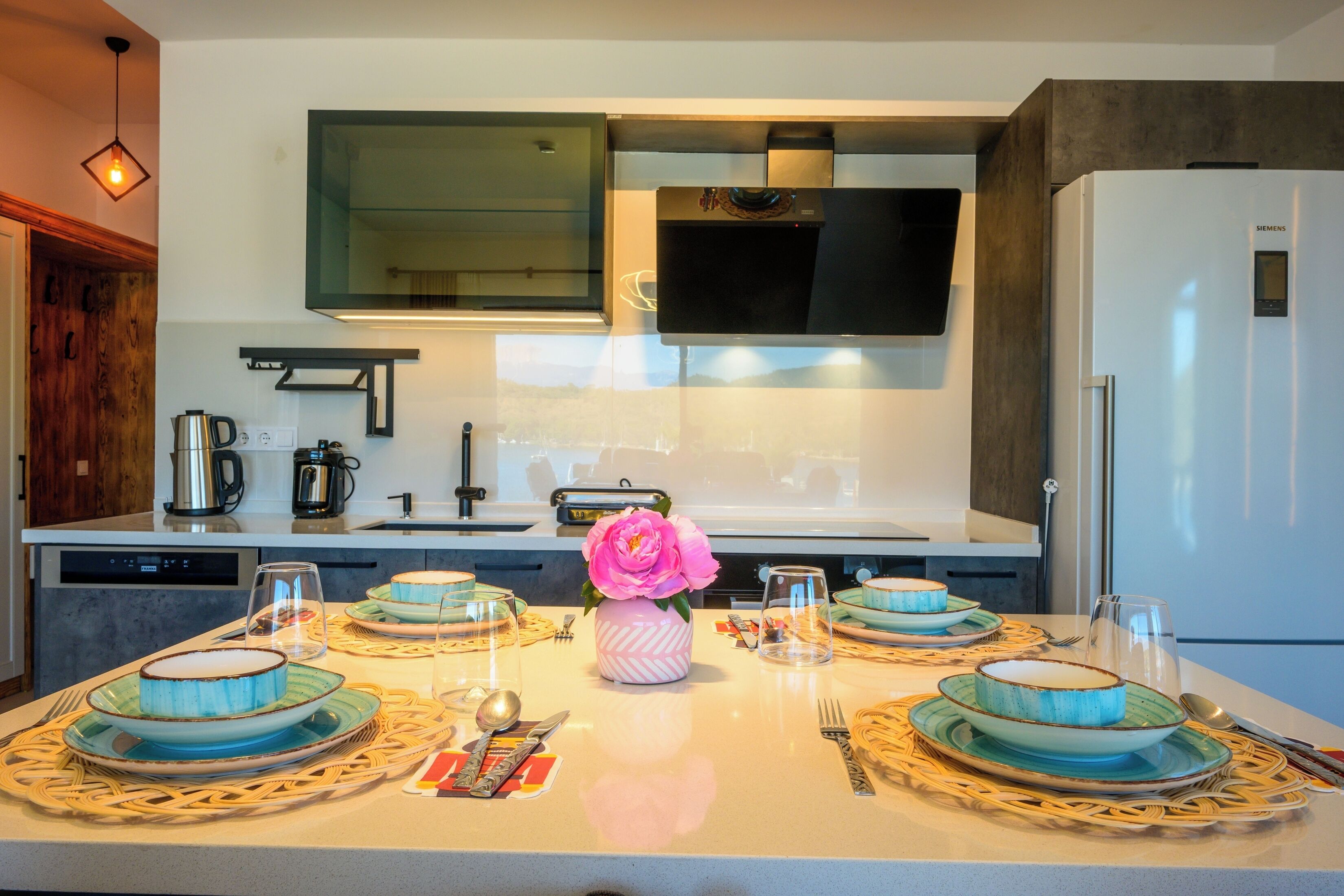 A modern kitchen with a dining table set for four, featuring blue and green plates and a pink flower centerpiece.