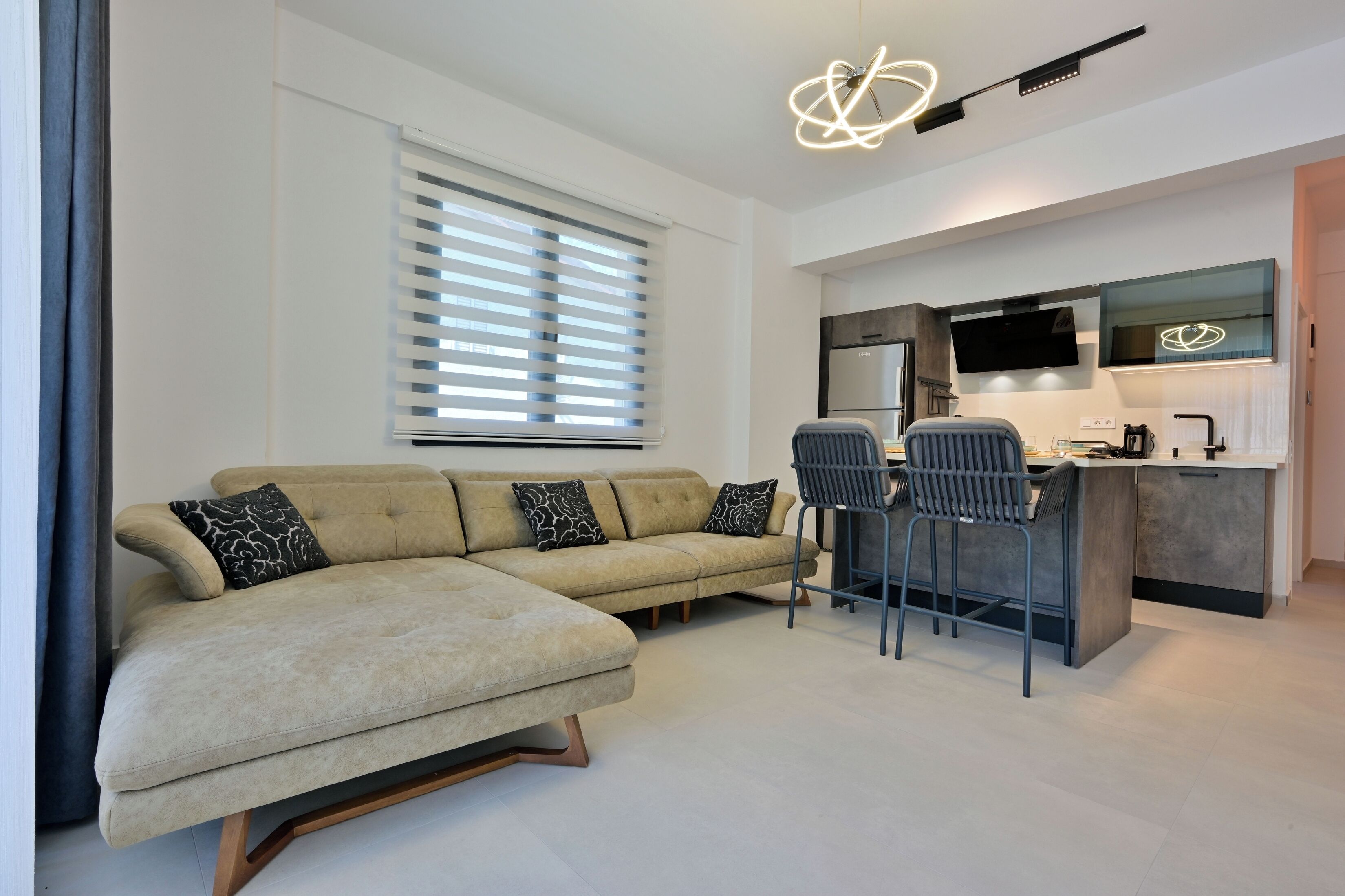 A modern living room with a sofa, two chairs, a kitchen area with a TV, and a unique ceiling light fixture.