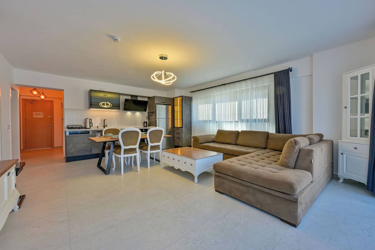 A modern living room with a beige sofa, a dining table with chairs, a kitchen area with a TV, and a large window with curtains.