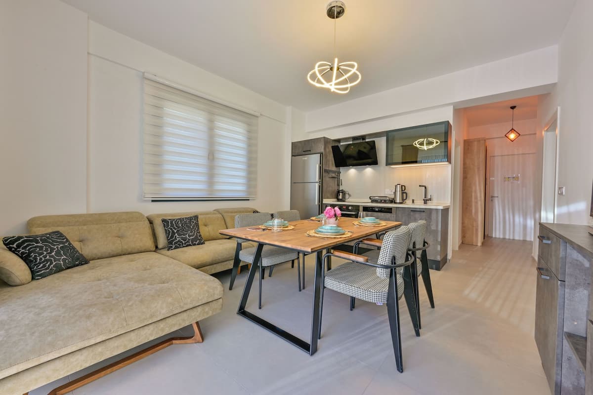 A modern living room with a sofa, dining table, and chairs. There is a kitchen area with a refrigerator, microwave, and sink.