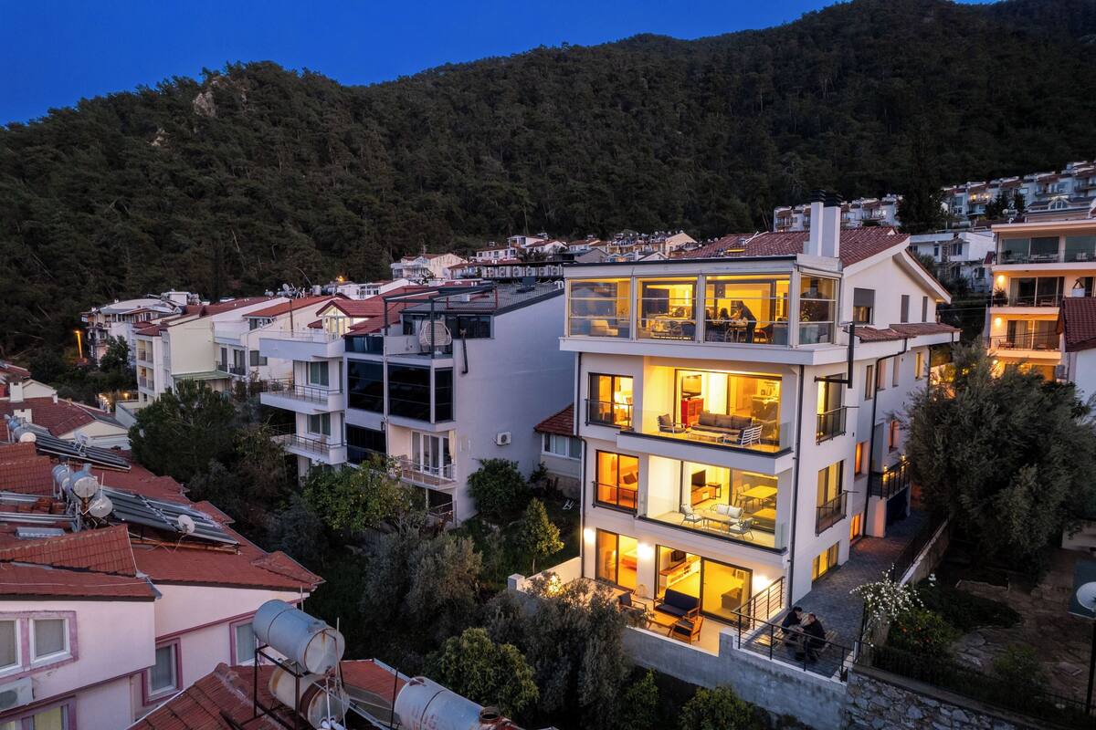 A multi-story residential building with balconies, large windows, and a view of a forested hillside.