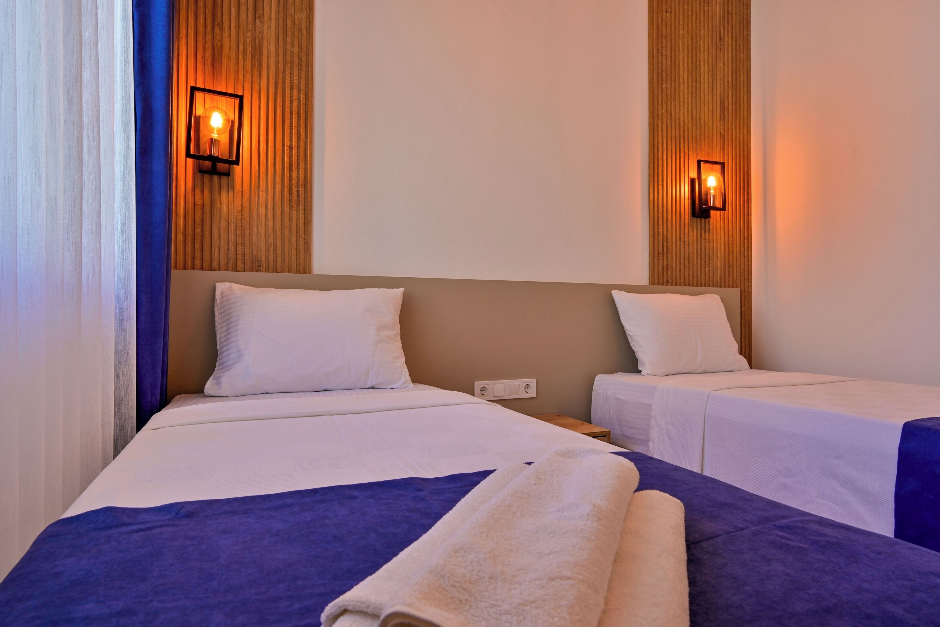 A hotel room with two beds, each with white linens and a blue accent. There are wall-mounted light fixtures on either side of the beds.