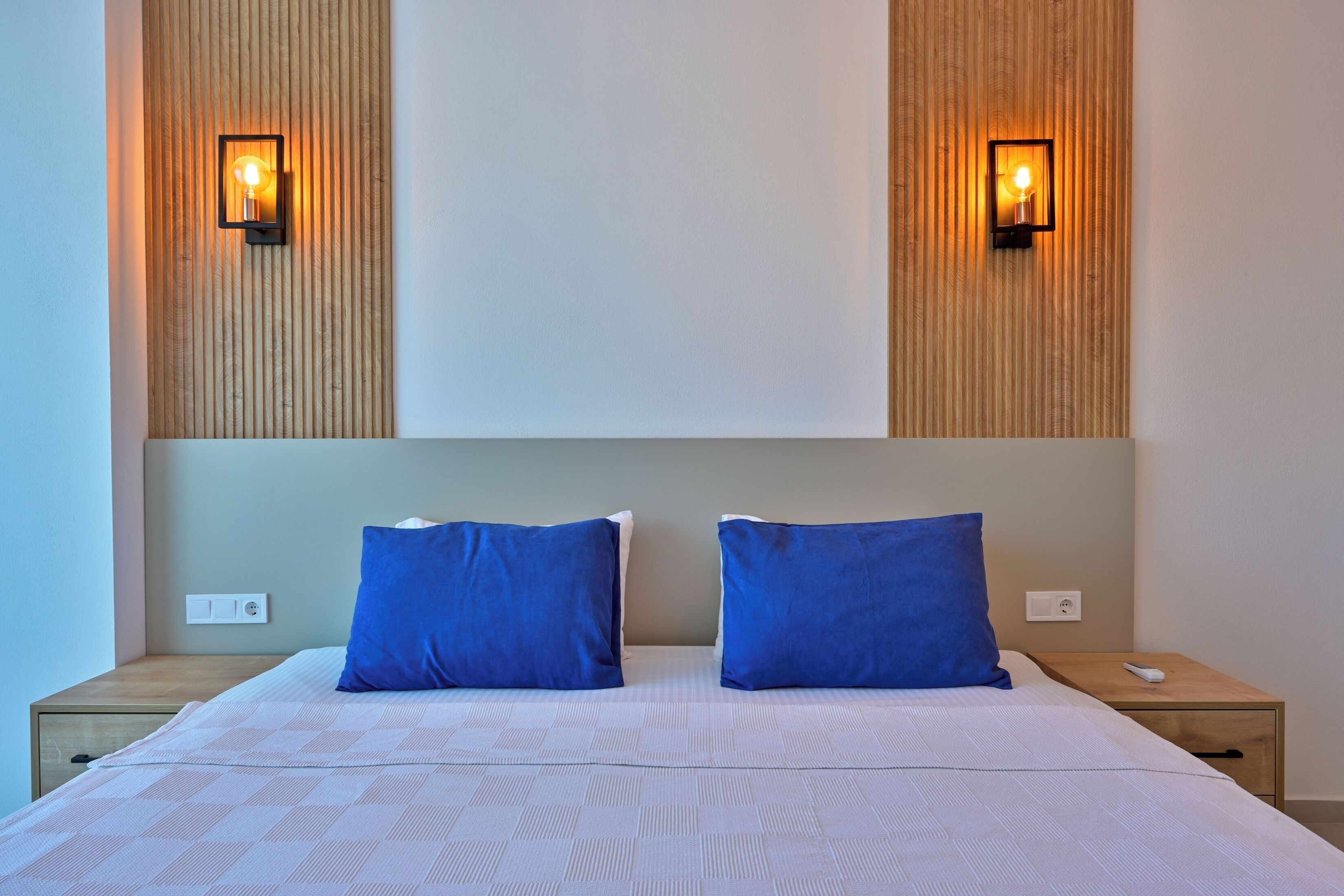 A neatly made double bed with two blue pillows, a wooden headboard, and two wall-mounted lights.