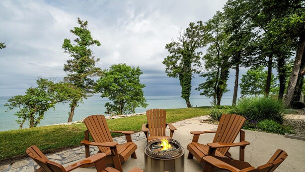 A circular seating arrangement with six Adirondack chairs around a central fire pit, overlooking a body of water and surrounded by trees. - (3 Nt Min) Lakefront Stunner Has It All! (Stevensville)