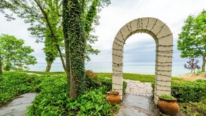 A stone archway with a pathway leading to a beach, surrounded by greenery and potted plants. - (3 Nt Min) Lakefront Stunner Has It All! (Stevensville)
