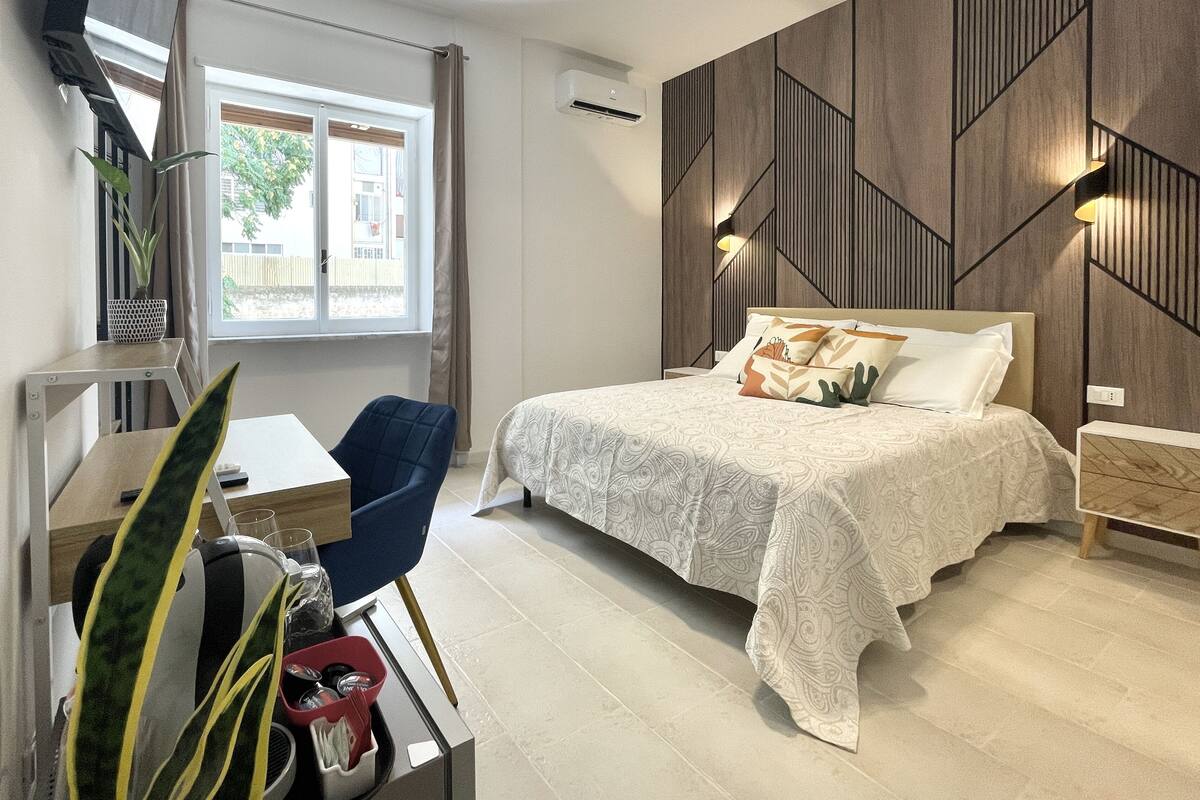 A modern bedroom with a bed, a desk, and a chair. There is a window with curtains, an air conditioning unit, and a wall-mounted light fixture.