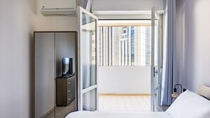 A modern hotel room with a bed, a wardrobe, a desk with a lamp, and a view of the city through large windows. - Casa Kenzo With Parking by Napoliapartments (Naples)