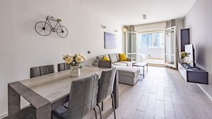 A modern living room with a dining area, grey sofas, a coffee table, and a mounted bicycle. - Casa Kenzo With Parking by Napoliapartments (Naples)