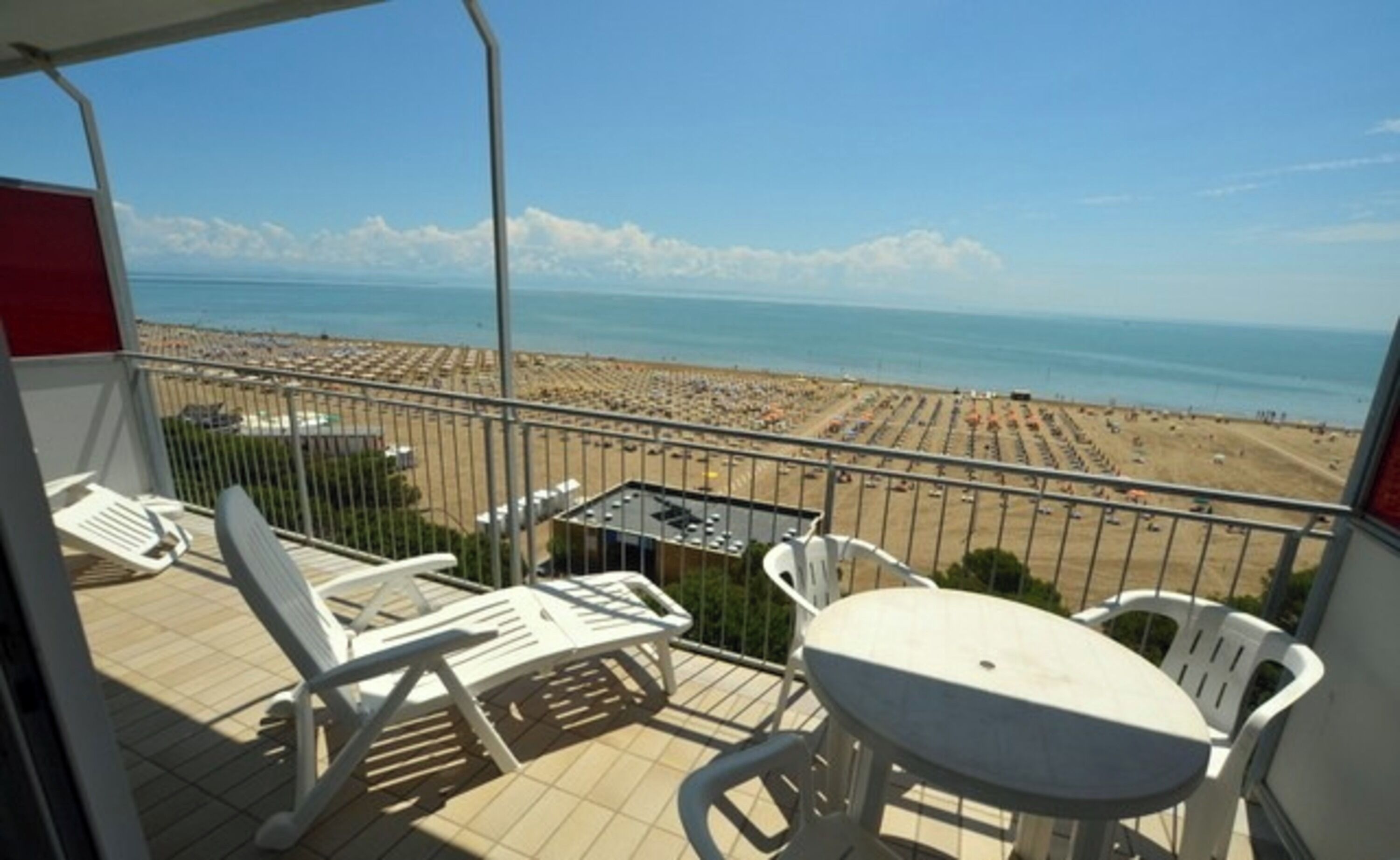 Varanda com cadeiras e mesa de plástico branco, com vista para uma praia com fileiras de guarda-sóis e pessoas.