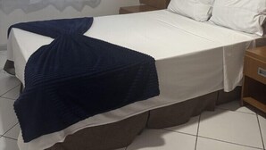 A neatly made bed with white linens and a blue blanket, a wooden nightstand with a lamp, and a window with sheer curtains. - Flat Aconchegante  (Campos)