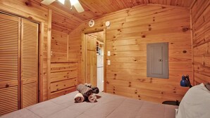 Ein Holzblockhaus-Zimmer mit einem Bett, einem kleinen Tisch mit Handtüchern, einem Ventilator und einer Tür zu einem anderen Raum.